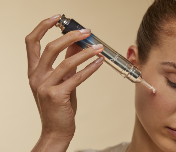 A person applies skincare serum under their eye using a metallic dropper syringe, showcasing innovative beauty packaging against a beige background. Only part of their face and hand are visible.