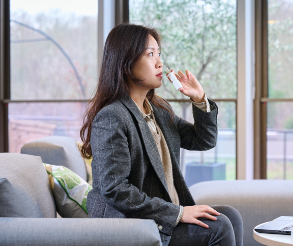 An Asian woman with long, dark hair sits in a modern living room, holding the APF Futurity™ recyclable nasal spray by Aptar Pharma close to her nose. She is wearing a grey checkered coat and is in the process of using the spray.