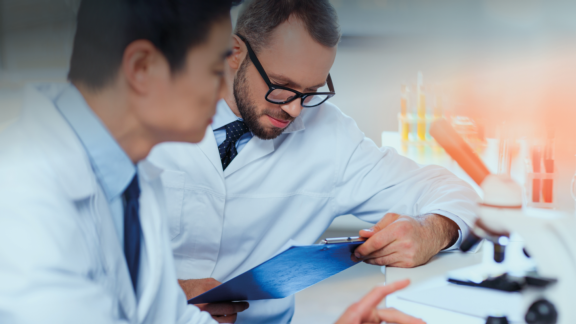 Two men sitting at laboratory benchtop in white lab coats review oral liquid product development data beside microscope.