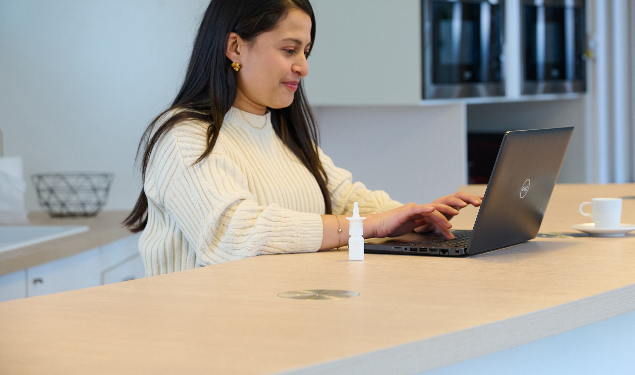 Woman with black hair in white sweater sitting at desk with laptop and APF futurity recyclable nasal spray.