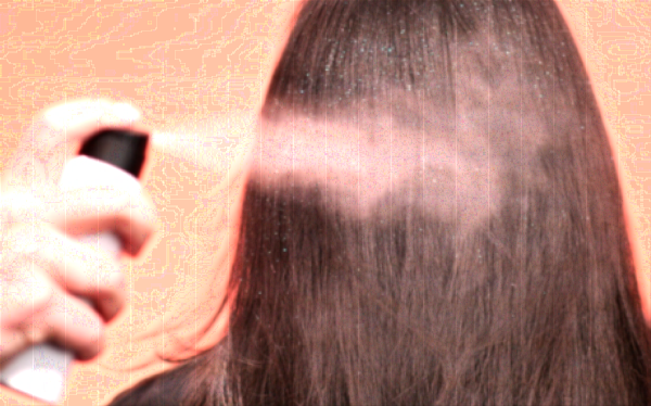 An image of a white aerosol spray can with a black actuator spraying dry shampoo and glitter on brown hair, depicting that a vertical slider aerosol valve is highly compatible with high powder formulations. The background is coral pink