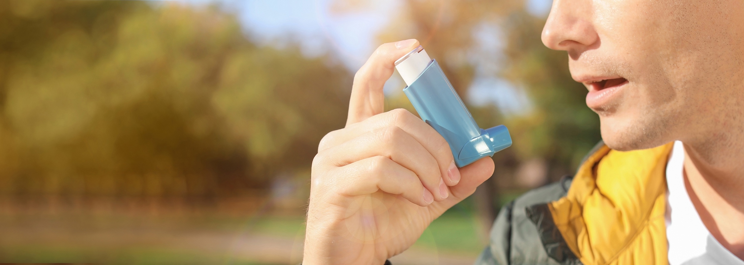 Man in green jacket in forested outdoor setting administering blue Aptar Pharma inhaler.