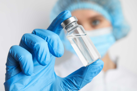 Aptar Pharma laboratory scientist wearing blue gloves and hairnet holds a capped glass vial filled with clear liquid.