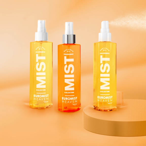 Three spray bottles labeled  EUROMIST HEAVEN stand on a peach-colored surface and background, with one bottle with a silver collar ,and one spraying a fine mist. The bottles are in shades of yellow and orange.